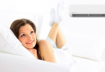 A woman lies comfortably on a white sofa, smiling and looking up. Relaxed with her head on a cushion, she enjoys the cool air from an efficient air conditioner—made possible by solar rebate savings.