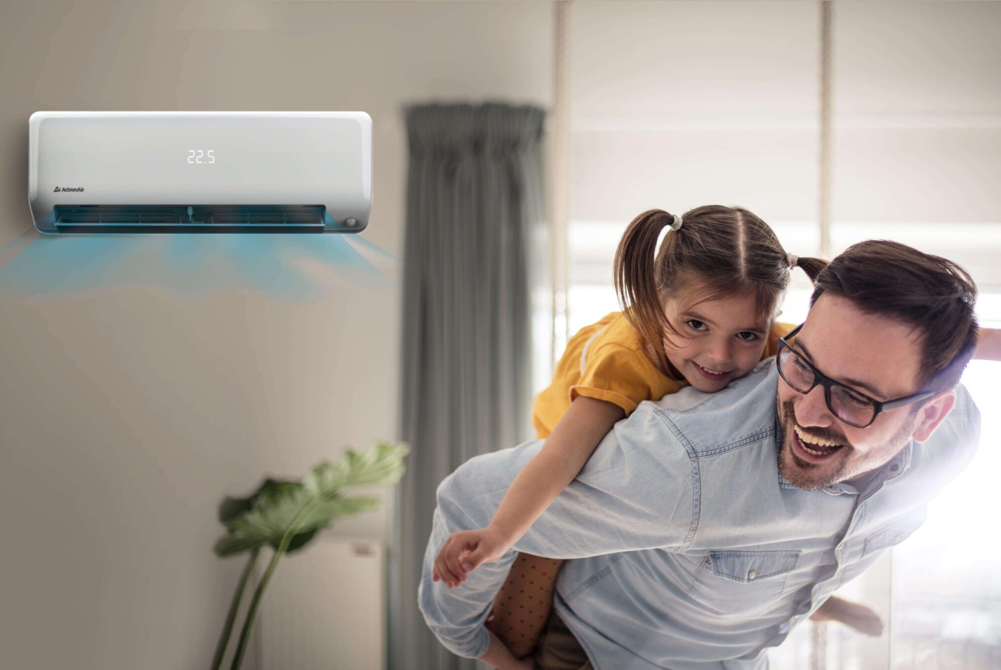 A man with glasses gives a piggyback to a smiling young girl in a bright living room. An air conditioner, possibly bought with an appliance rebate, displays 22.5°C. A leafy plant and curtained window are in the background.