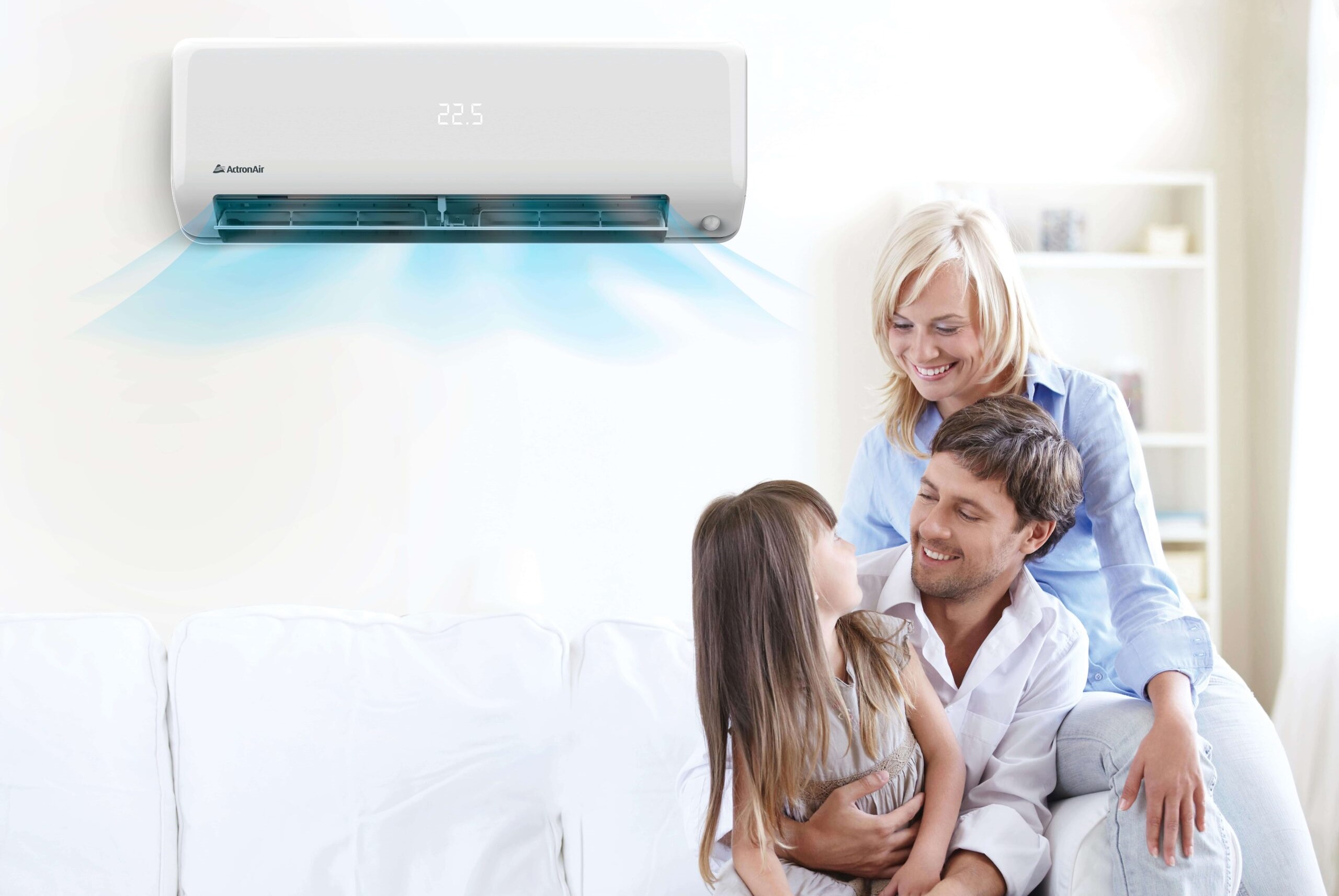 A smiling family of three sits on a white sofa under a wall-mounted air conditioner, which displays 22.5°C and releases cool blue air—showcasing the comfort possible with an appliance rebate in their bright, cosy room.