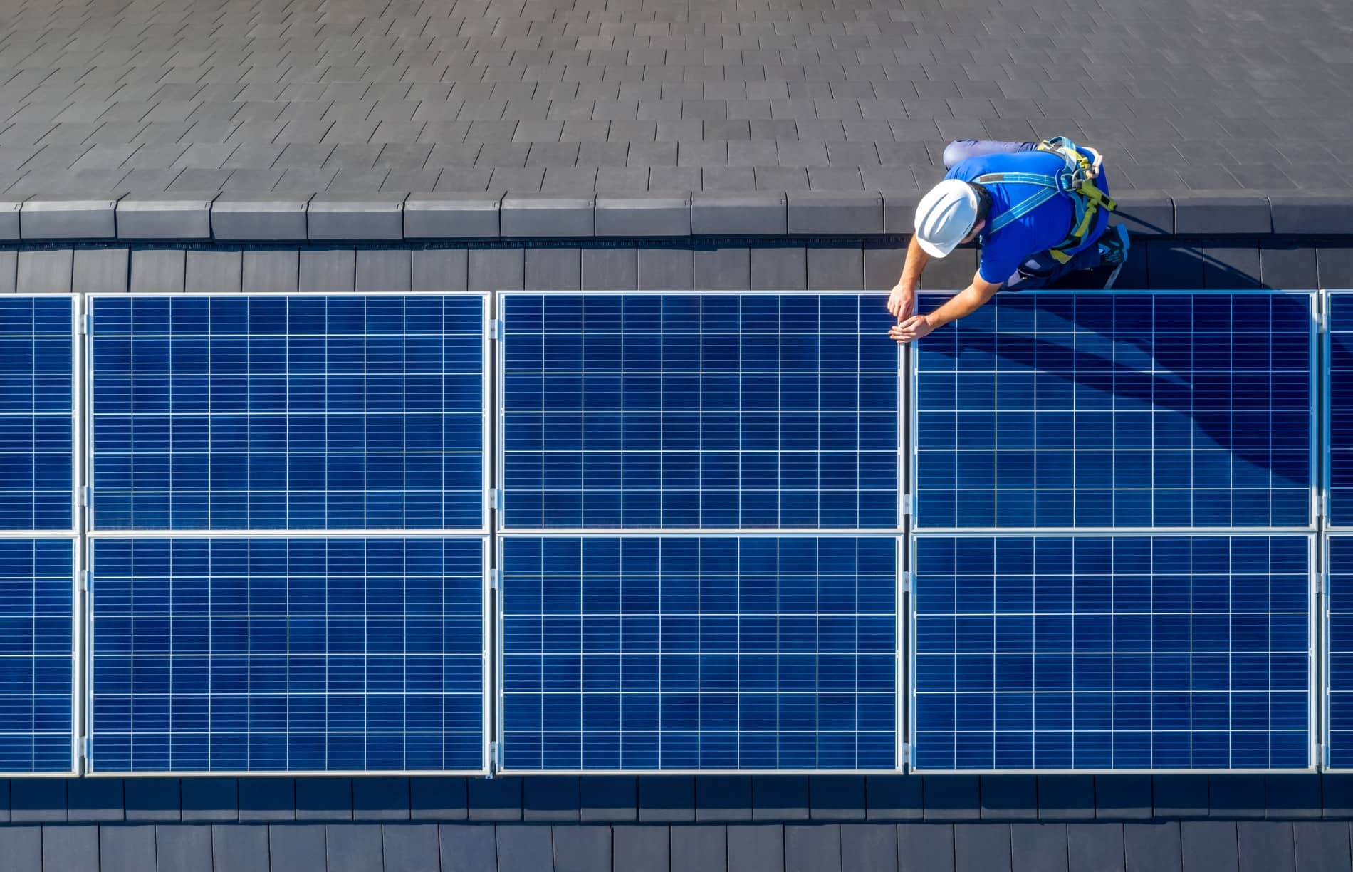 A worker in safety gear installs or inspects blue solar panels on a sloped, tiled roof, viewed from above—taking advantage of a solar rebate through MAC Trade Services.