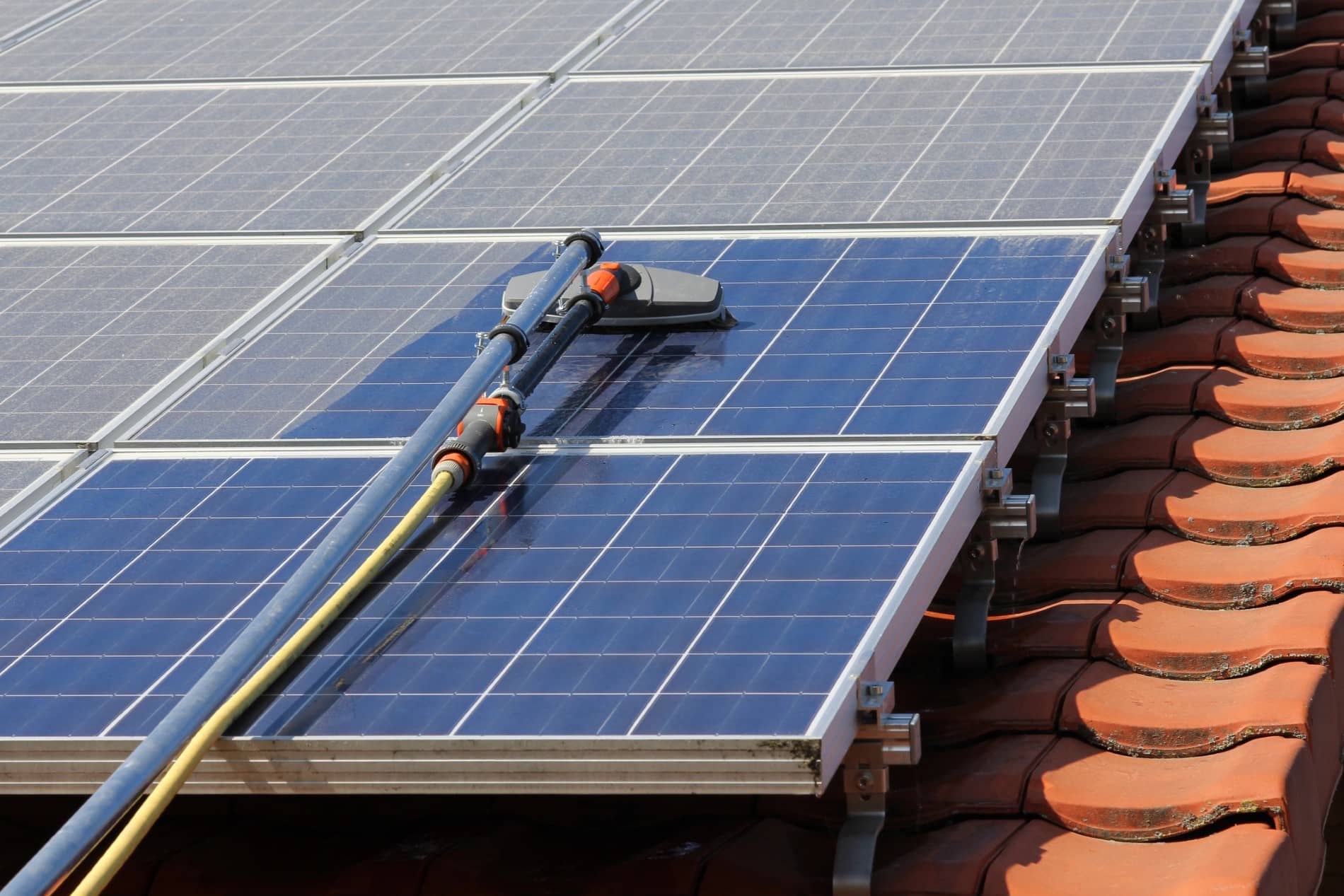 A close-up view of solar panels on a tiled roof being cleaned with a brush attached to a hose, spraying water over the panel surfaces—professionally maintained by Mac Trade Services to help homeowners maximise their solar rebate.
