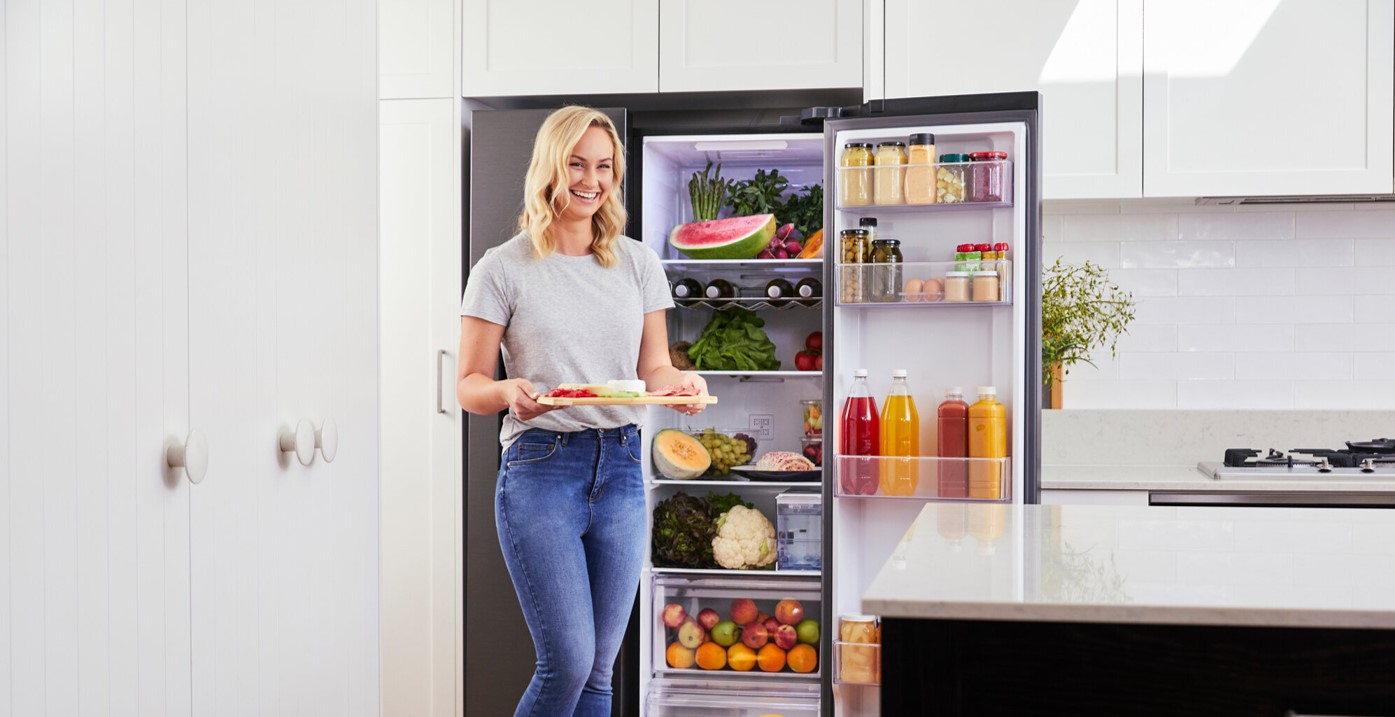 A woman smiles whilst standing in a modern kitchen, holding a plate of food. Behind her, an open fridge is stocked with groceries—showcasing fresh choices powered by solar rebate savings.