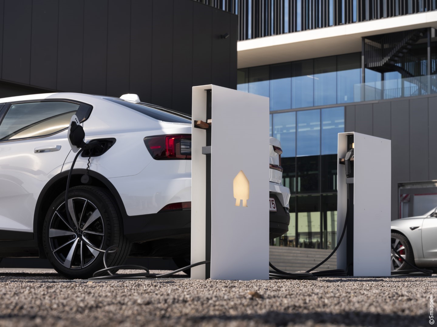 A white electric car is parked and plugged into a modern charging station outside a contemporary glass building, showcasing eco-friendly travel. Appliance rebate programmes encourage more drivers to embrace sustainable choices like this.