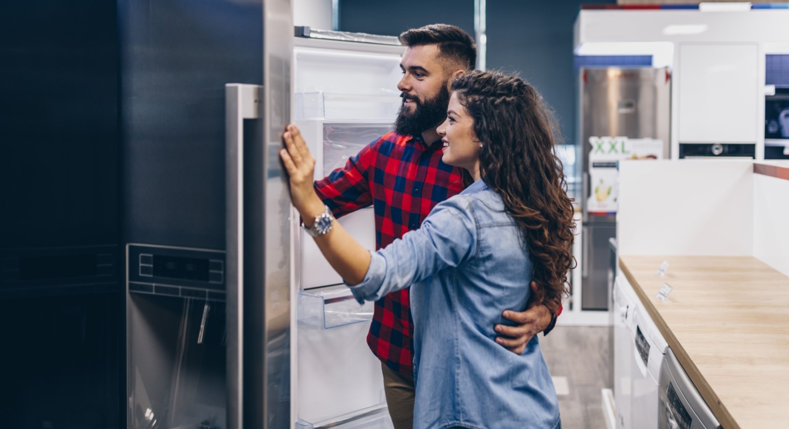 A smiling couple stands in an appliance shop, looking inside an open fridge. The man is embracing the woman as they examine the fridge together, discussing possible appliance rebate offers among other appliances.