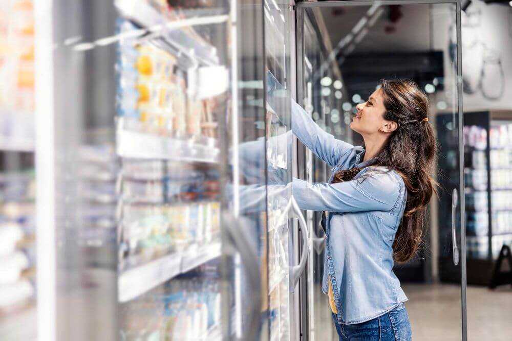 A woman wearing a light denim jacket is reaching into a refrigerated section at a supermarket, possibly comparing items eligible for an appliance refund, surrounded by shelves filled with various products.
