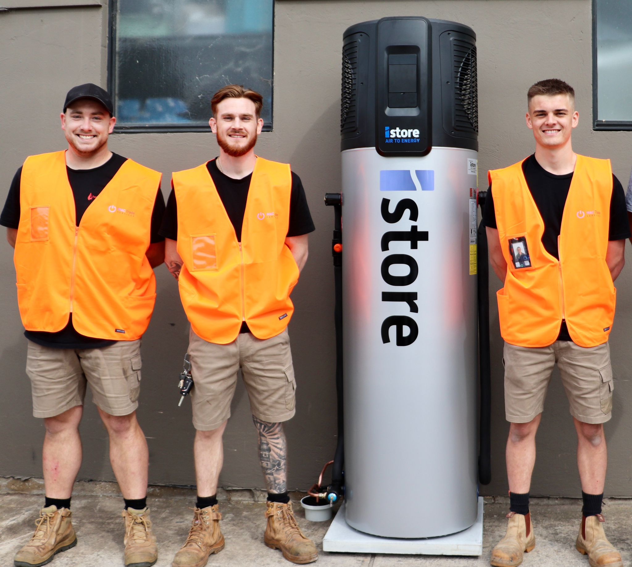 Three smiling workers in orange safety vests and shorts stand beside a large, cylindrical iStore water heater outside a building with grey walls, highlighting the benefits of an appliance rebate through Mac Trade Services.