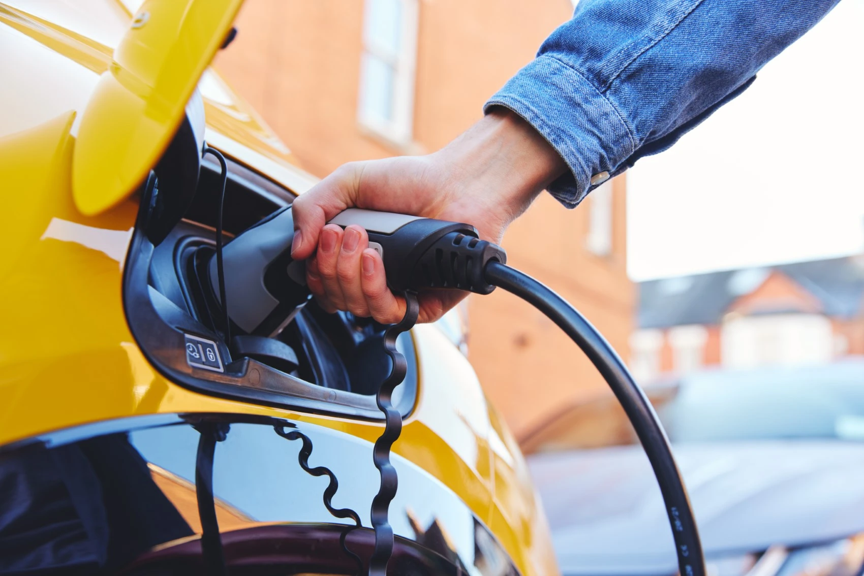 A person’s hand plugs a charging cable into a yellow electric car, with buildings and another car visible in the blurred background—perfect for those looking to maximise savings with a battery rebate.