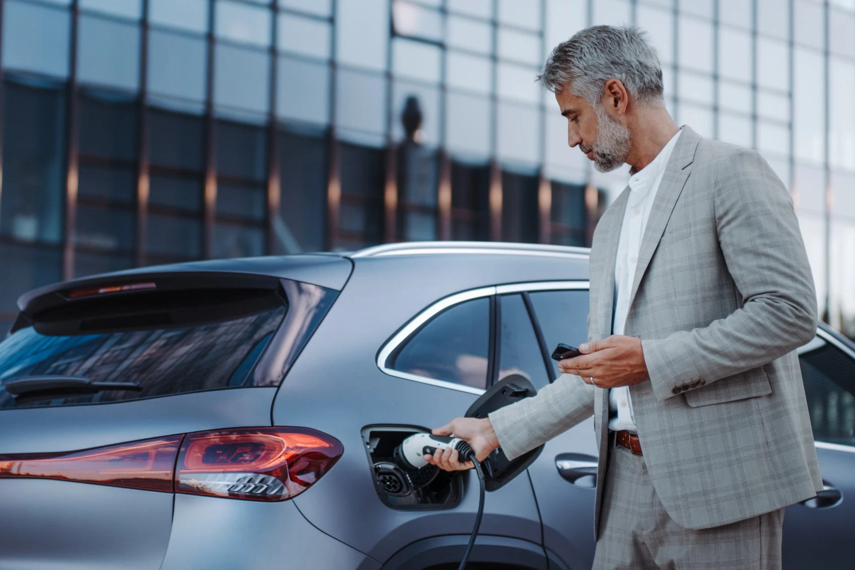 A man in a grey suit stands next to a silver electric car, plugging in a charging cable. He holds a mobile phone in his other hand, possibly checking his solar rebate information, as a modern glass building rises behind him.