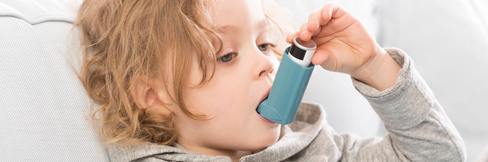 A young child with curly hair uses an inhaler whilst sitting on a light-coloured sofa, wearing a grey hoodie—whilst their parent reads about a solar rebate nearby.