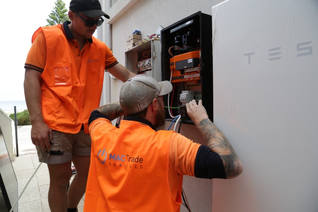 Two workers in orange vests install or repair electrical wiring in a control panel beside a Tesla Powerwall battery, possibly as part of a battery rebate upgrade. Tools and electrical components are visible.