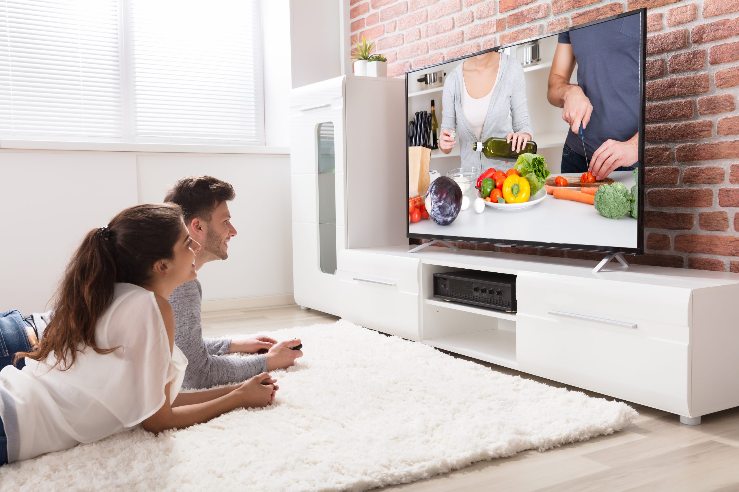 Happy Young Couple Sitting On Sofa Watching Recipe On Television At Home