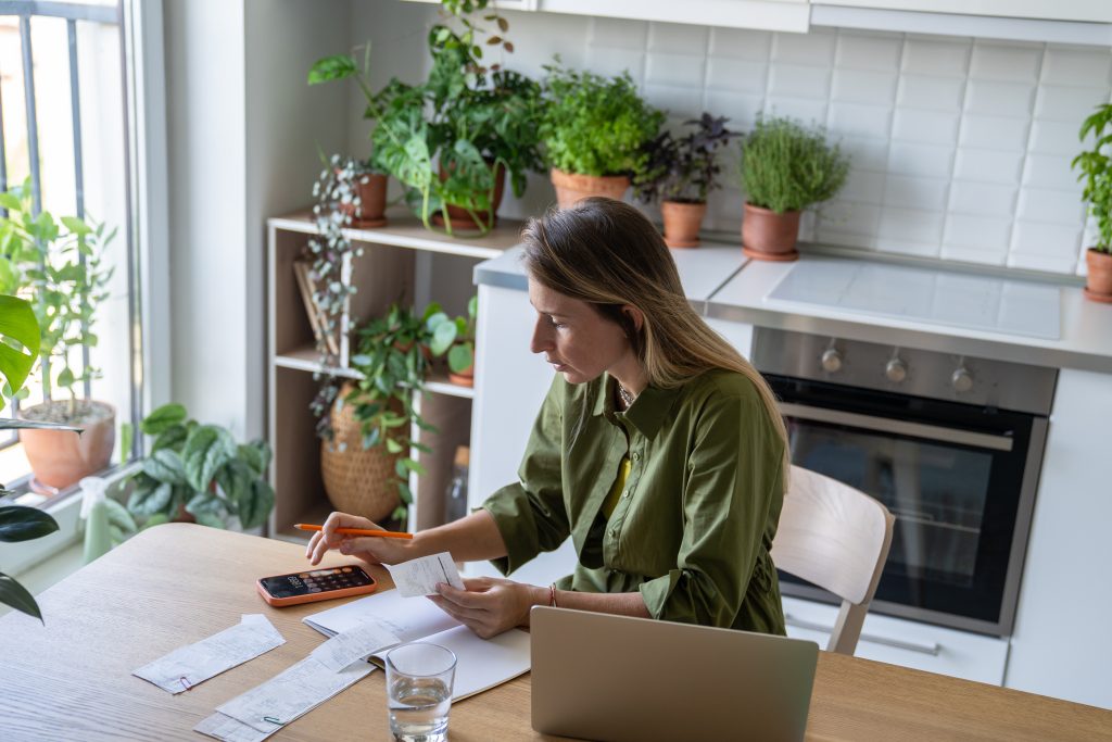 Woman sitting at a kitchen table calculating household expenses with receipts, calculator, and laptop surrounded by indoor plants