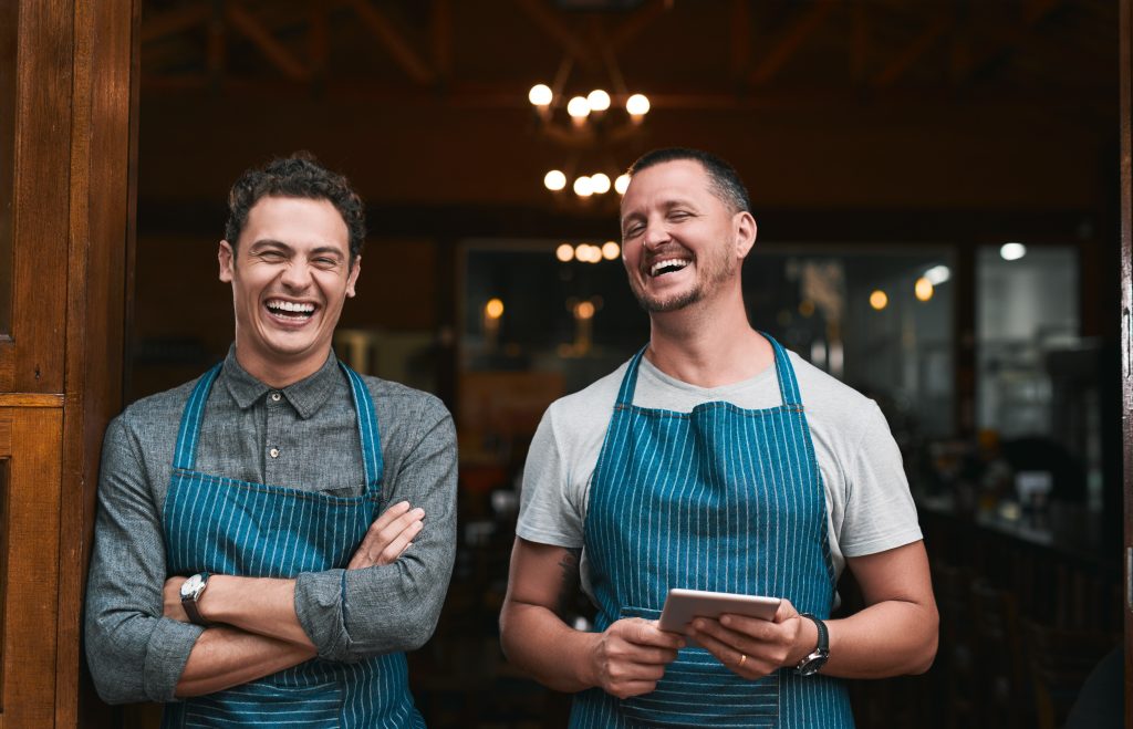 Two cheerful small business owners in aprons standing at their café entrance, using a digital tablet to manage their business efficiently