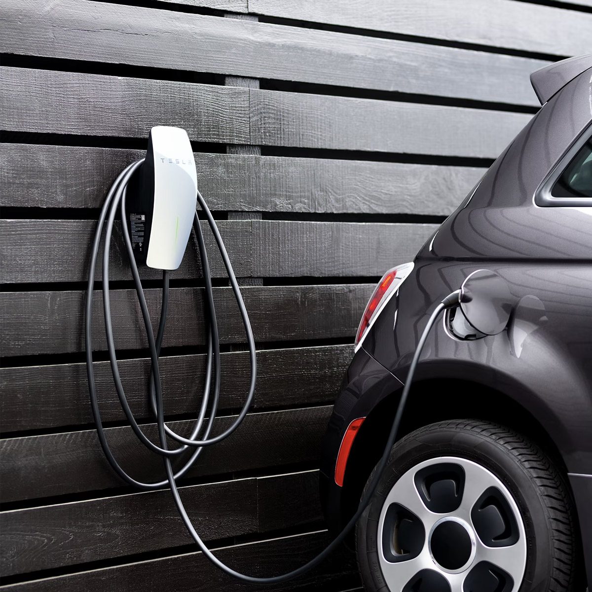 A black electric car is plugged into a Tesla wall charger on a dark wooden wall, with charging cables hanging neatly—perfect for taking advantage of a battery rebate to power your vehicle sustainably.