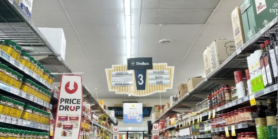 A supermarket aisle with shelves stocked with spices, sauces, and packaged foods. A sign overhead marks aisle 3 for Asian, Mexican, and Indian foods. A price reduction sign for taco shells is visible on the left alongside an appliance rebate display.
