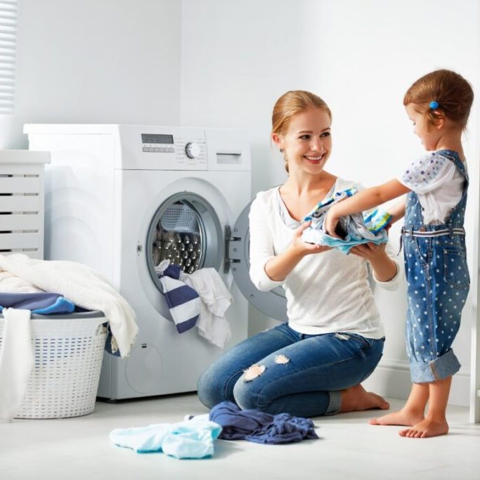 A smiling woman kneels by a front-loading washing machine, helping a young girl hand her clothes. Laundry baskets and detergent sit in a bright, tidy utility room—perfect for families interested in appliance rebate savings from Mac Trade Services.