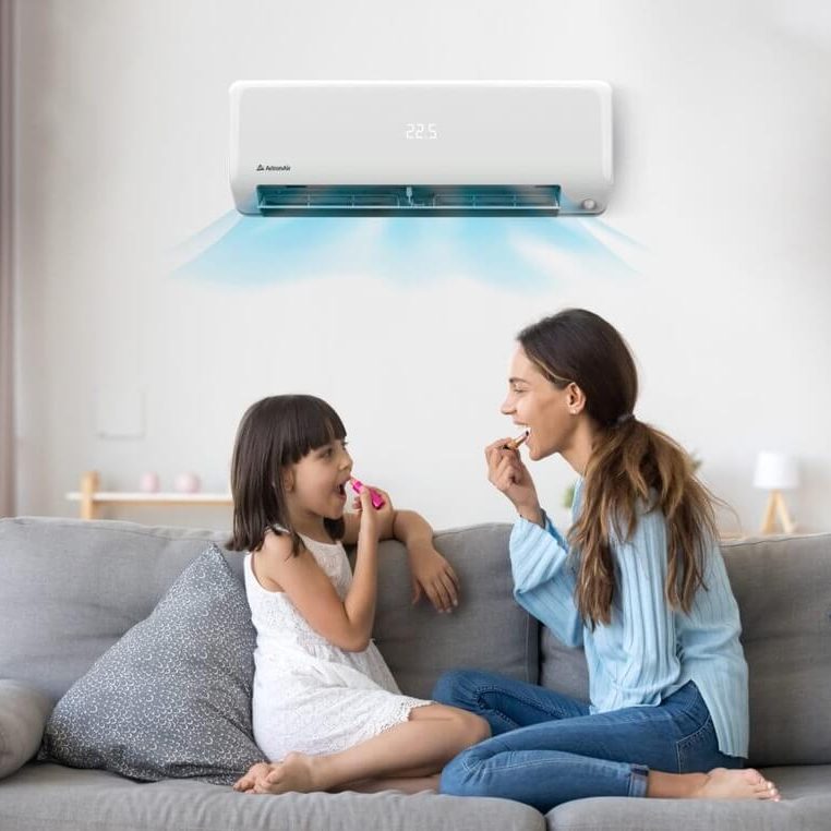 A woman and a young girl sit on a grey sofa, smiling and eating snacks as a wall-mounted air conditioner—eligible for an appliance rebate—blows cool air into the room.