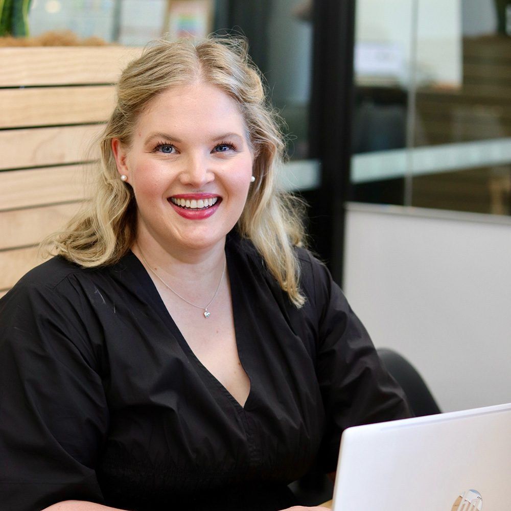 A smiling woman with blonde hair sits at a table, wearing a black blouse and working on a laptop in a modern office setting, researching appliance rebate options.