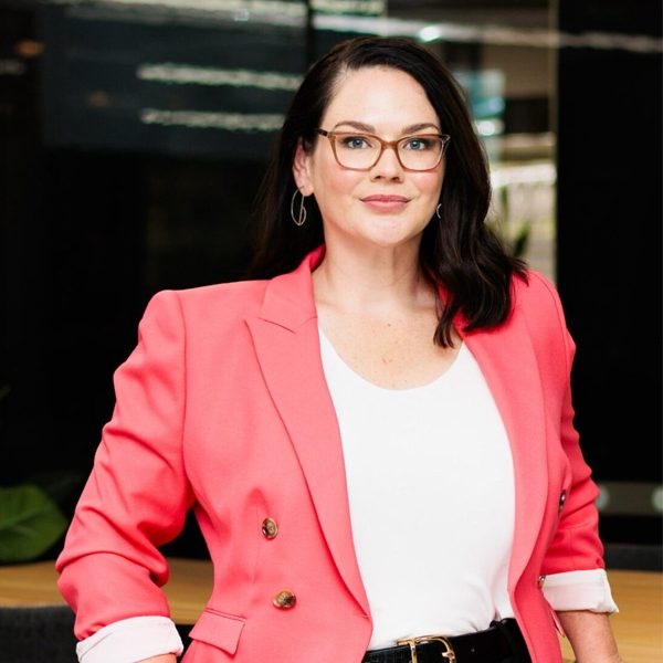 A woman with dark hair and glasses, wearing a coral blazer over a white top, stands confidently with one hand on her hip in a modern indoor setting, ready to discuss solar rebates and how you can benefit from government grants.